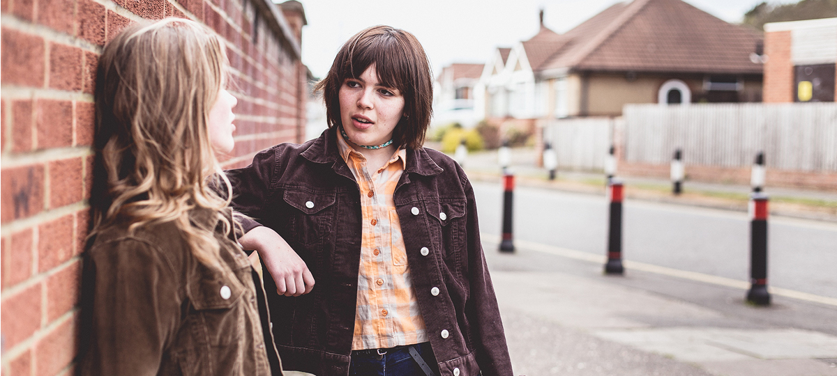 Leaning against a brick wall two teenagers have a conversation