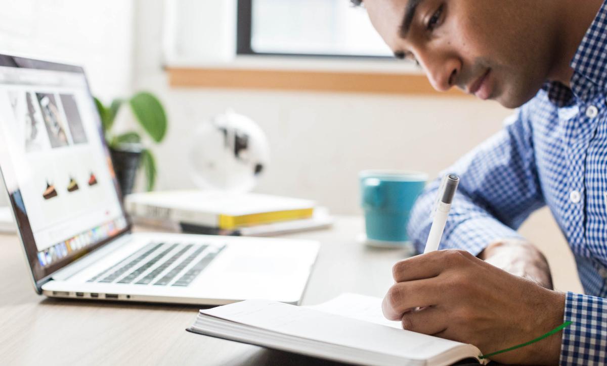 Man in shirt writing on paper on desk with laptop 