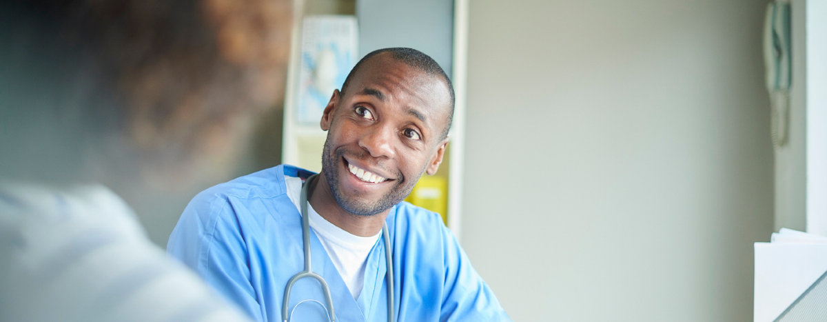 Male hospital doctor sat at a desk and making notes while talking to a female patient