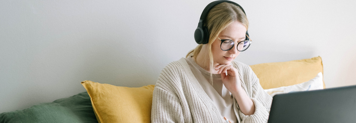 A woman sits on a coach working on a laptop with a pair of headphones on.