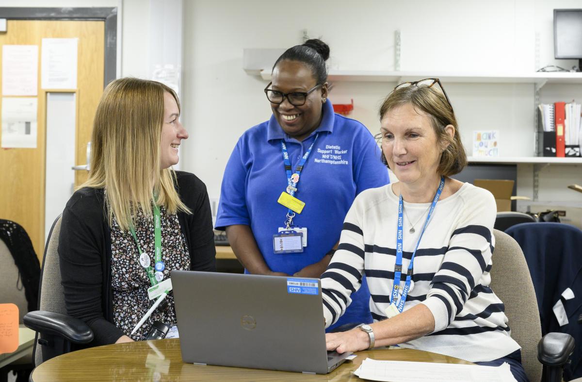 A photograph of three female members of NHFT staff smiling around a laptop computer