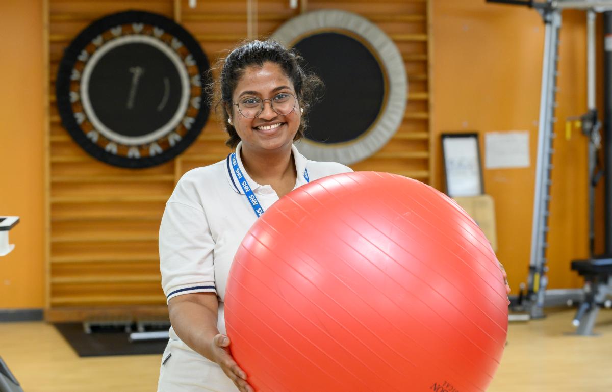 A female physiotherapist smiling and holding up an exercise ball in a gym setting