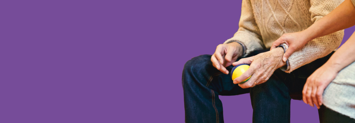 Older man holds a ball as a person checks he is okay against a purple background