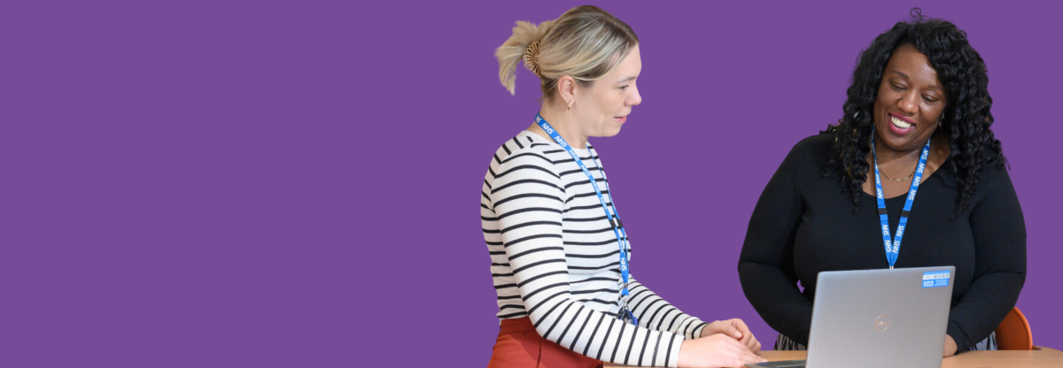 Two female members of staff talking in front of a laptop against a purple background