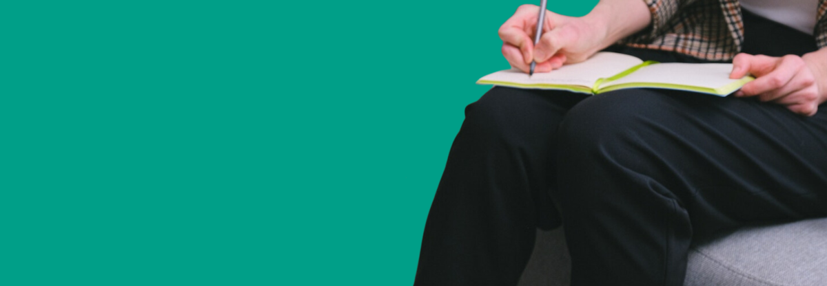 A close-up of a person writing in a notebook on their lap against a green background