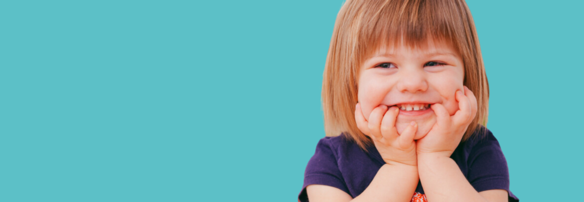 Toddler sits at a table laughing against a teal background