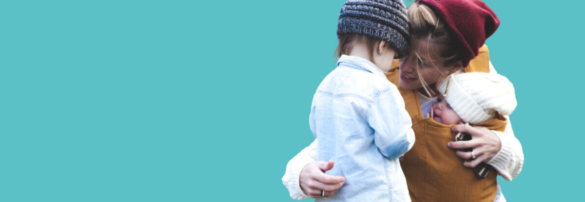 A parent kneels on the floor to comfort a toddler whilst holding a newborn baby against a teal background
