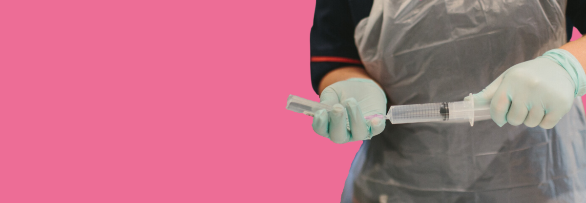 A close-up of a female nurse filling a syringe while wearing PPE against a pink background